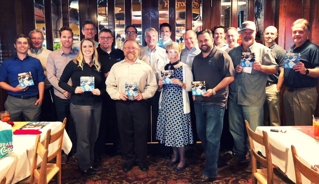 Members of the Capital Advisors group stand together in front of a wall with wooden and mirrored panels. They smile and hold up brochures from IDT Expo.
